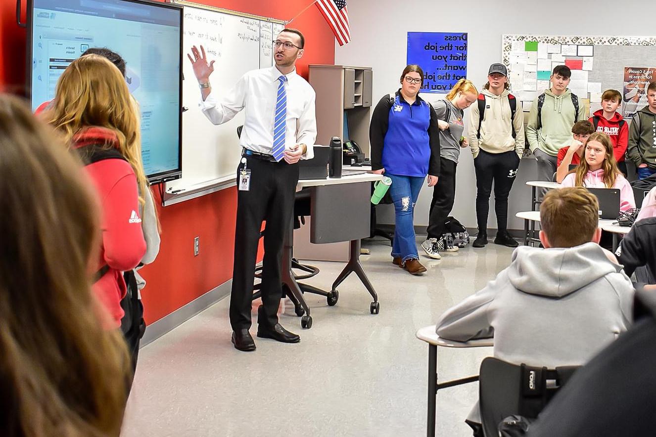 A man in a shirt and tie presenting around a group of students.